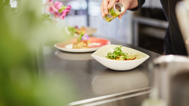 A cook seasons a dish in a bowl with herbs from a small tin.