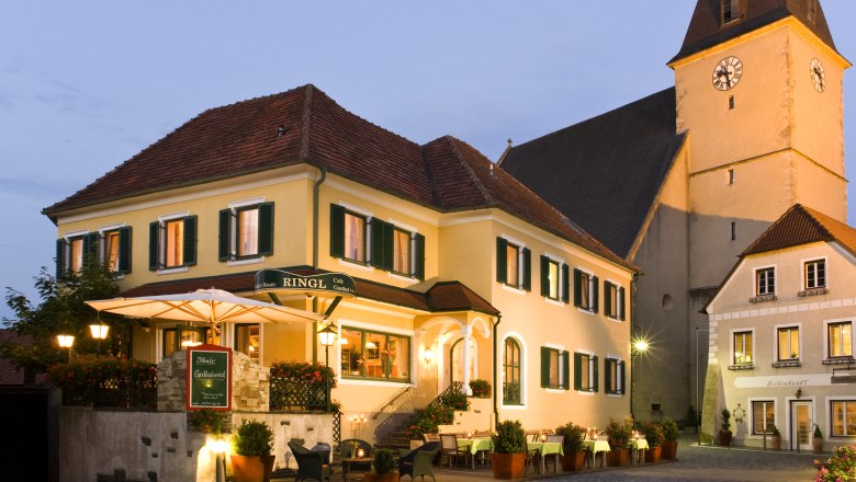 A traditional inn with a yellow fa&ccedil;ade and green shutters, illuminated at dusk, next to a church.