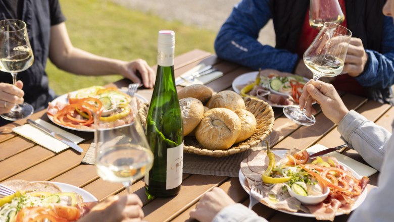 People enjoy wine and cold food outdoors at a wooden table.