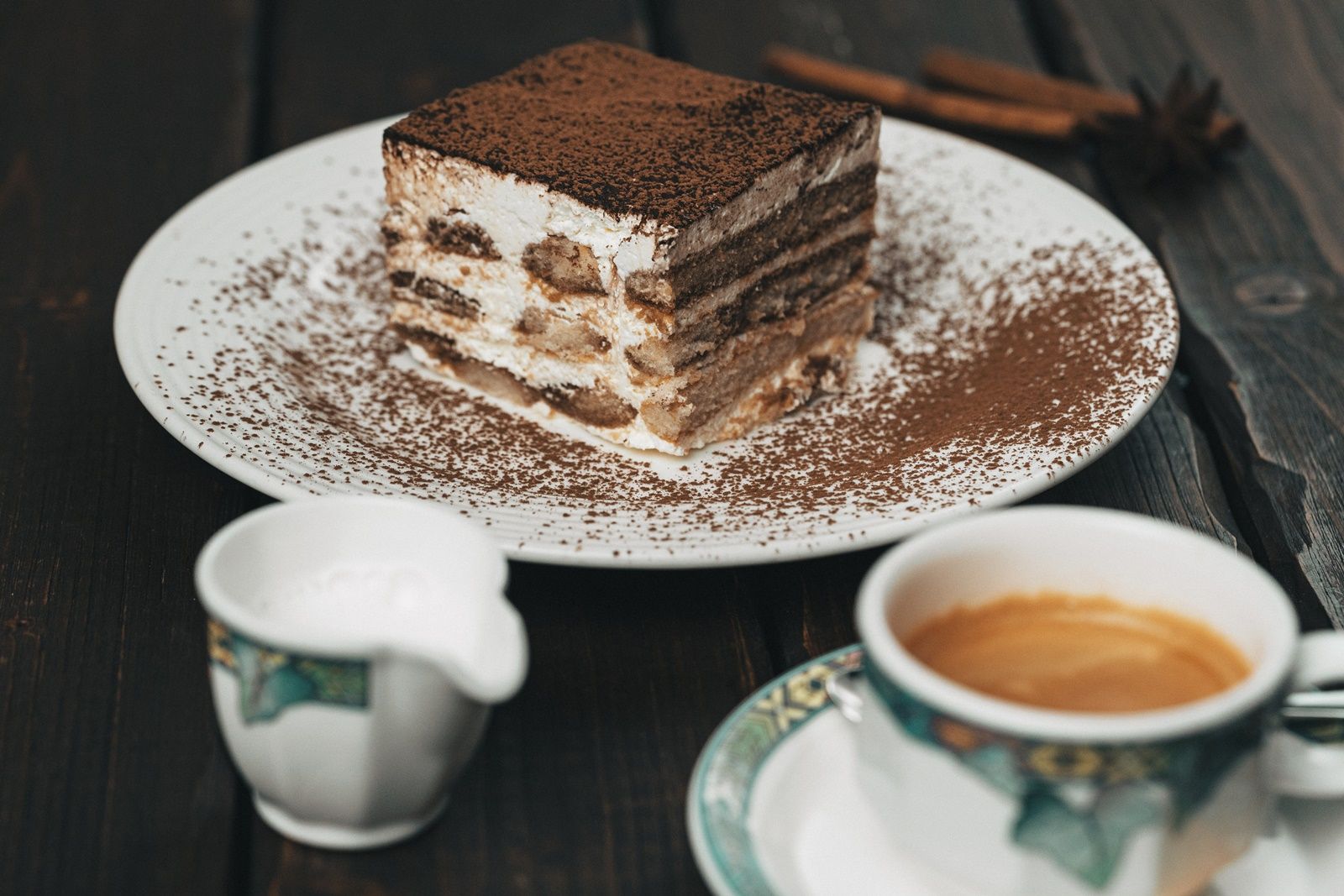 A plate with a slice of tiramisu, a cup of coffee and a milk jug next to it on a wooden table.