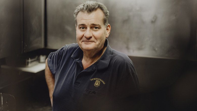 Man in a dark polo shirt with the "Dorfschenke" logo in a kitchen.