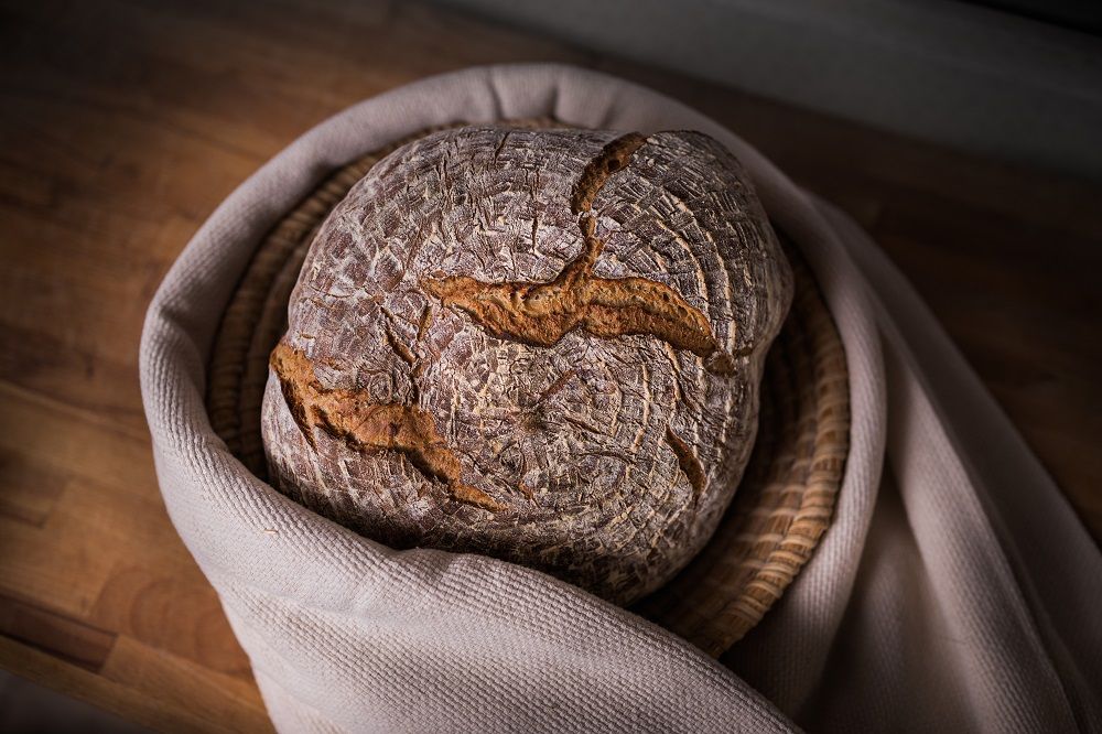 A loaf of bread in a basket on a wooden table, surrounded by a cloth.