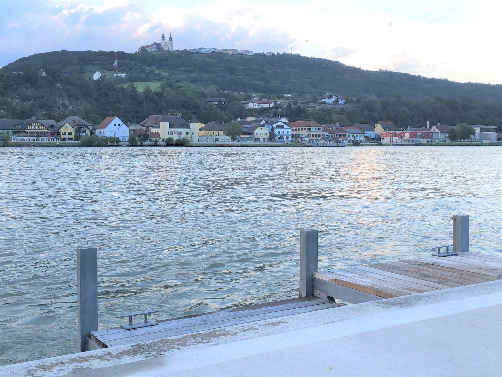 View from a landing stage of the river and colorful houses on the opposite bank, with a church on a hill in the background.