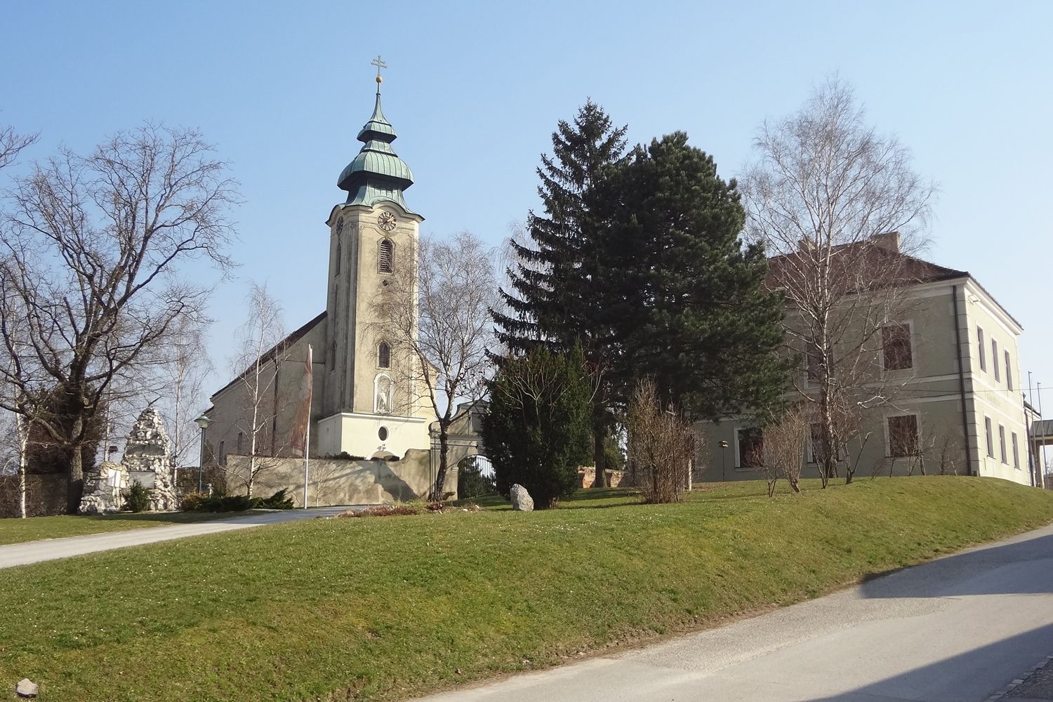 Church with tower and adjoining building in Gnadendorf, surrounded by trees and lawn.