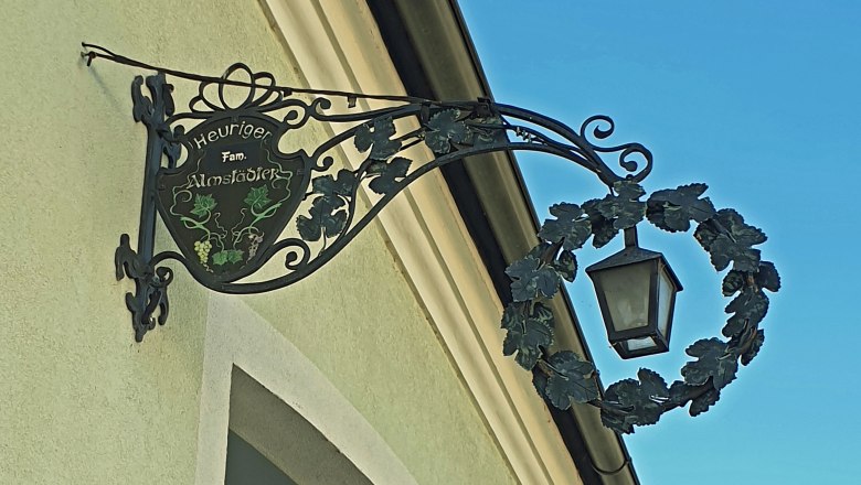 Metal sign with lantern on a house wall, labeled 'Heuriger Fam. Almst&auml;dter'.
