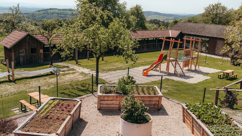 A playground with a slide and climbing frame next to raised beds in a garden with wooden huts in the background.