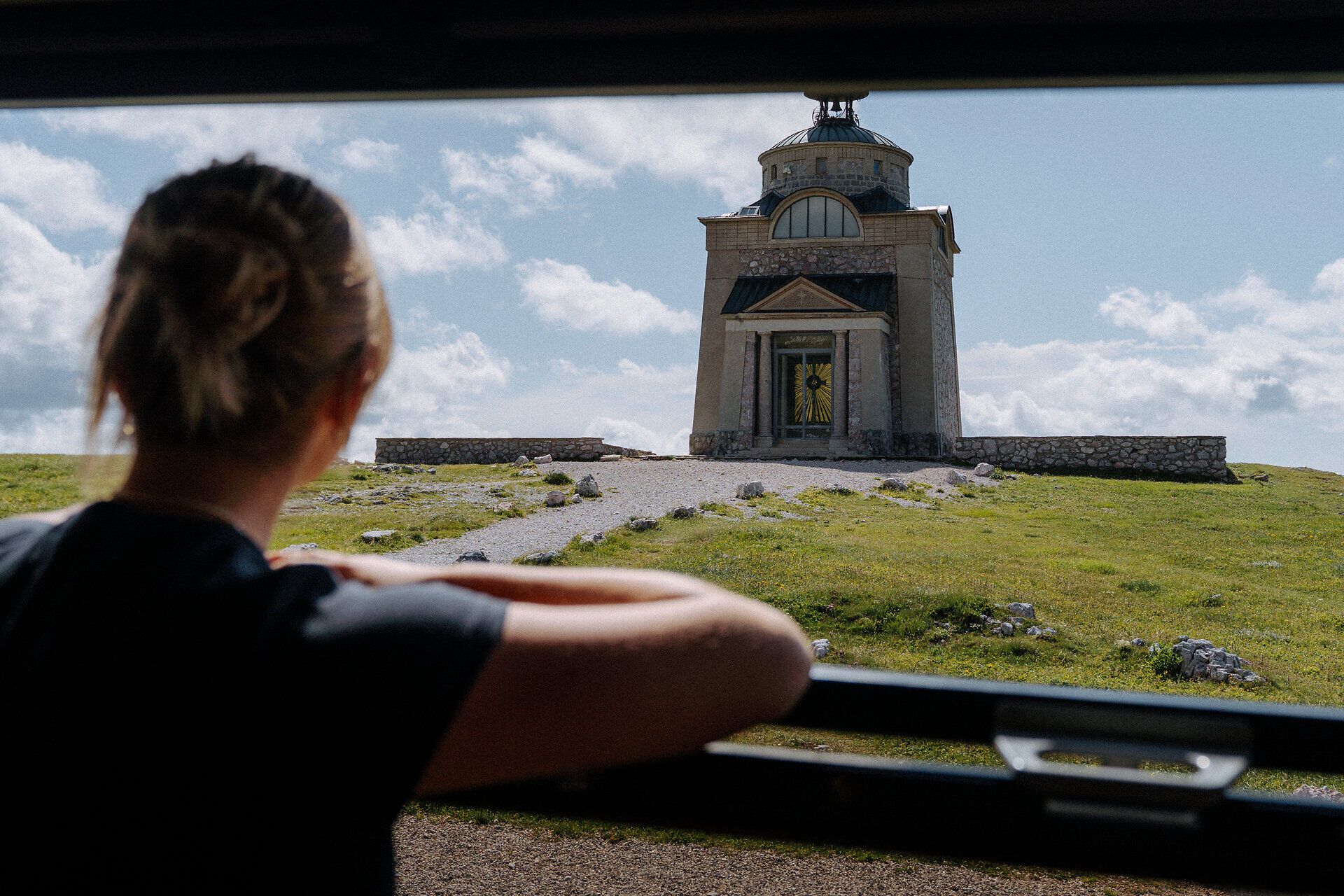 A woman looks out from the Schneebergbahn at the Elisabethkircherl on the Hochschneeberg. A gravel path leads to the church, which is surrounded by a green meadow, creating an atmosphere of exploration and historical curiosity.