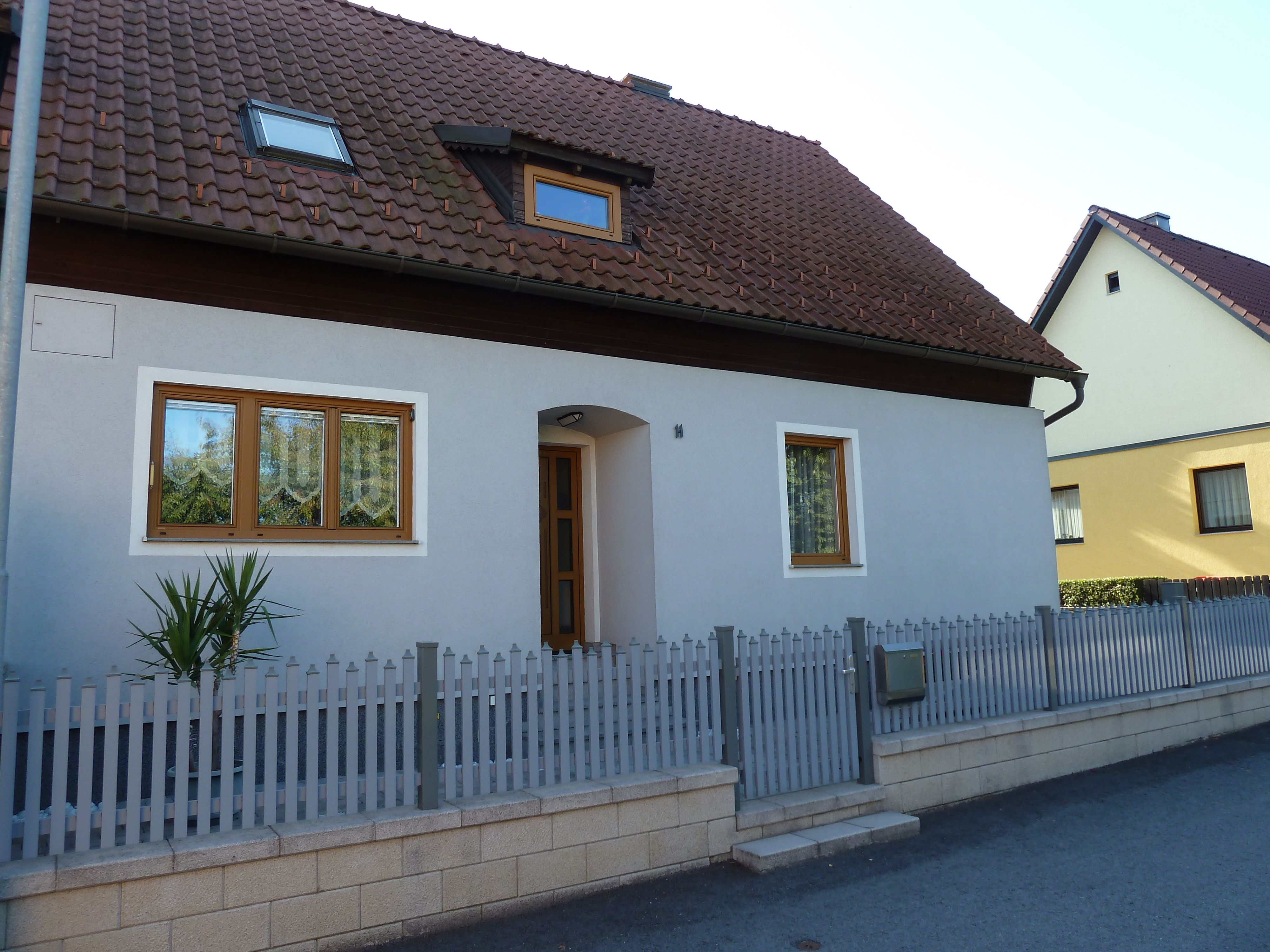 A detached house with a gray façade, brown roof and wooden doors. A small fence surrounds the property.