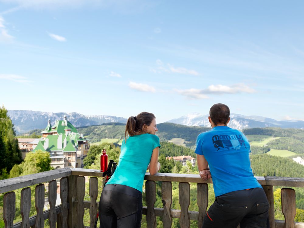 Two people stand on a balcony with a view of the Südbahnhotel and the surrounding mountains.
