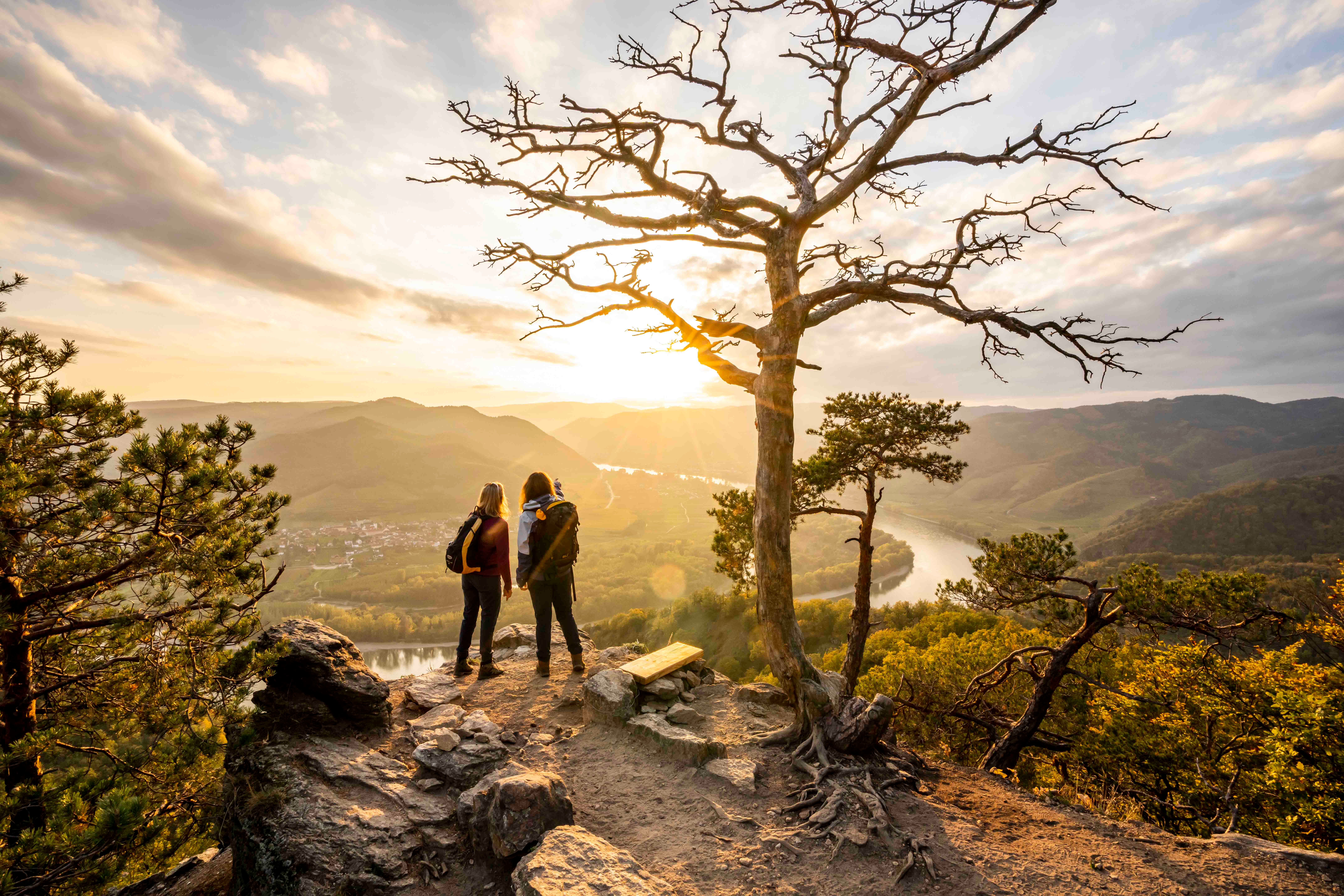 Two people stand on a rock with a view of a river landscape at sunset.