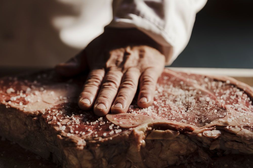 Close-up of a hand sprinkling coarse salt on a large piece of raw meat.