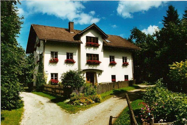 A traditional white house with red shutters and flower boxes, surrounded by trees and a garden, under a blue sky.