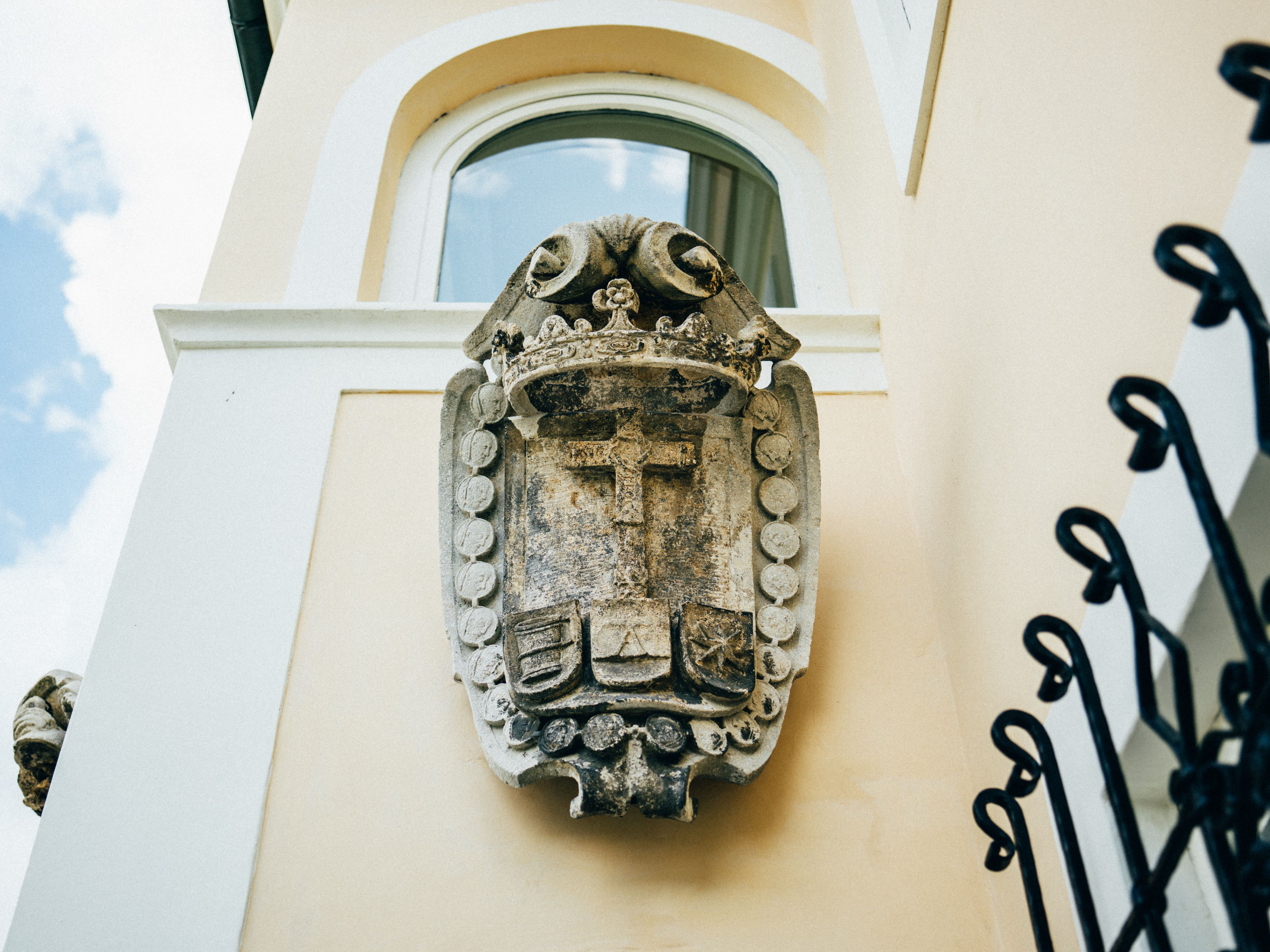 Stone coat of arms on a yellow house wall with window and blue sky in the background.