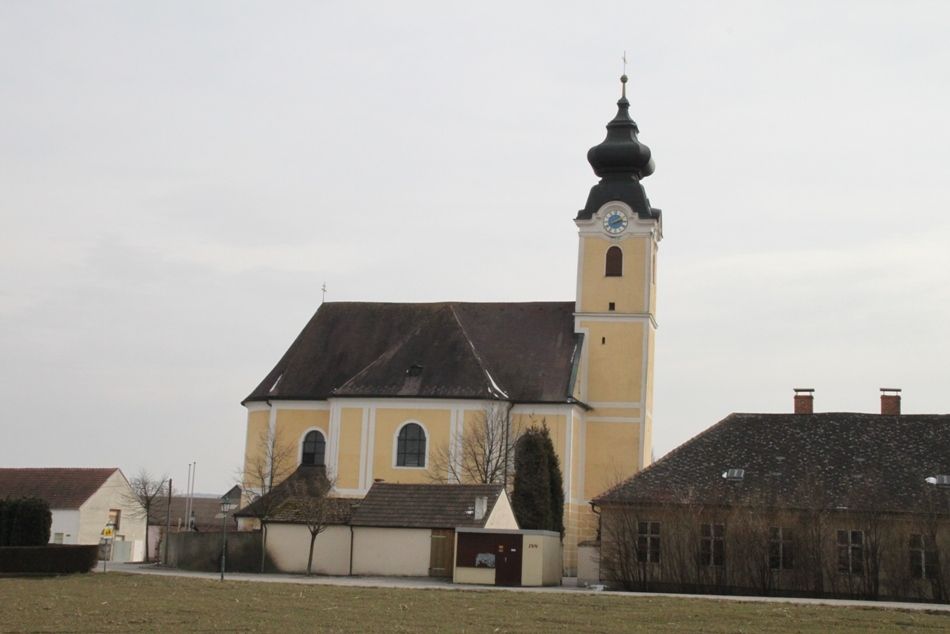 Yellow church with tower and clock in Langenrohr, surrounded by buildings and trees.