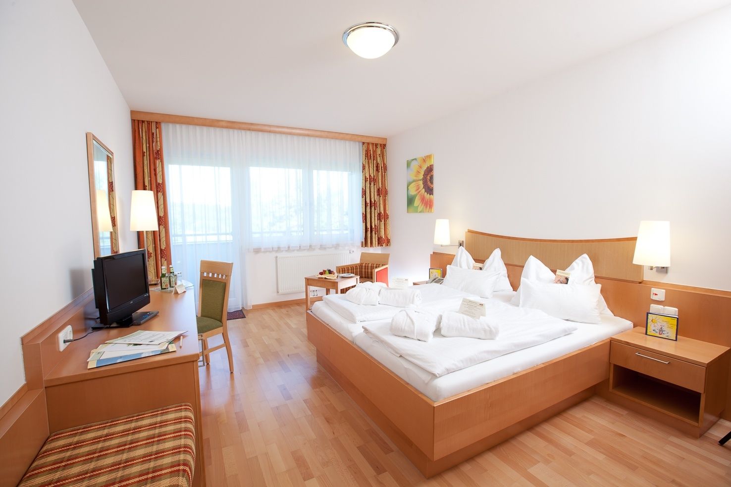 Bright hotel room with double bed, desk, TV and large windows.