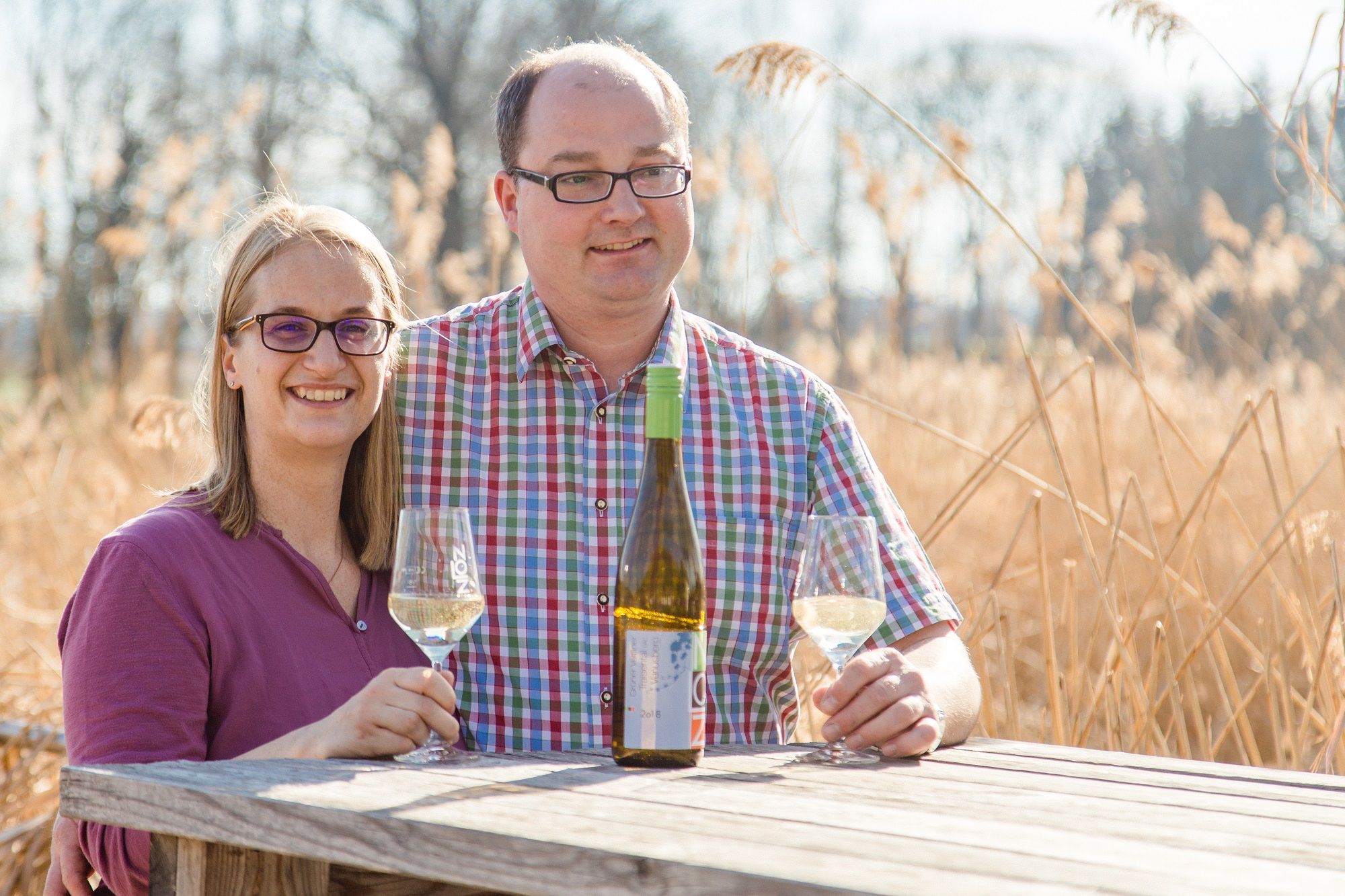 A man and a woman are sitting at a wooden table outside with wine glasses and a bottle of wine.