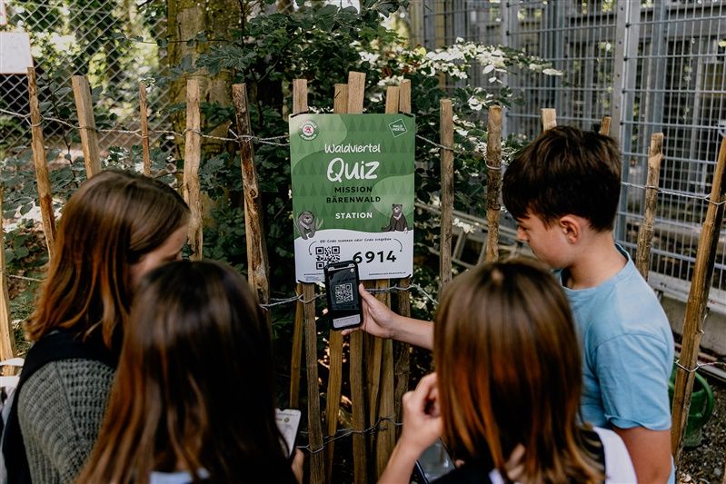 Children stand in front of a sign with the words 'Waldviertel Quiz Mission Bärenwald Station' and scan a QR code with a smartphone.