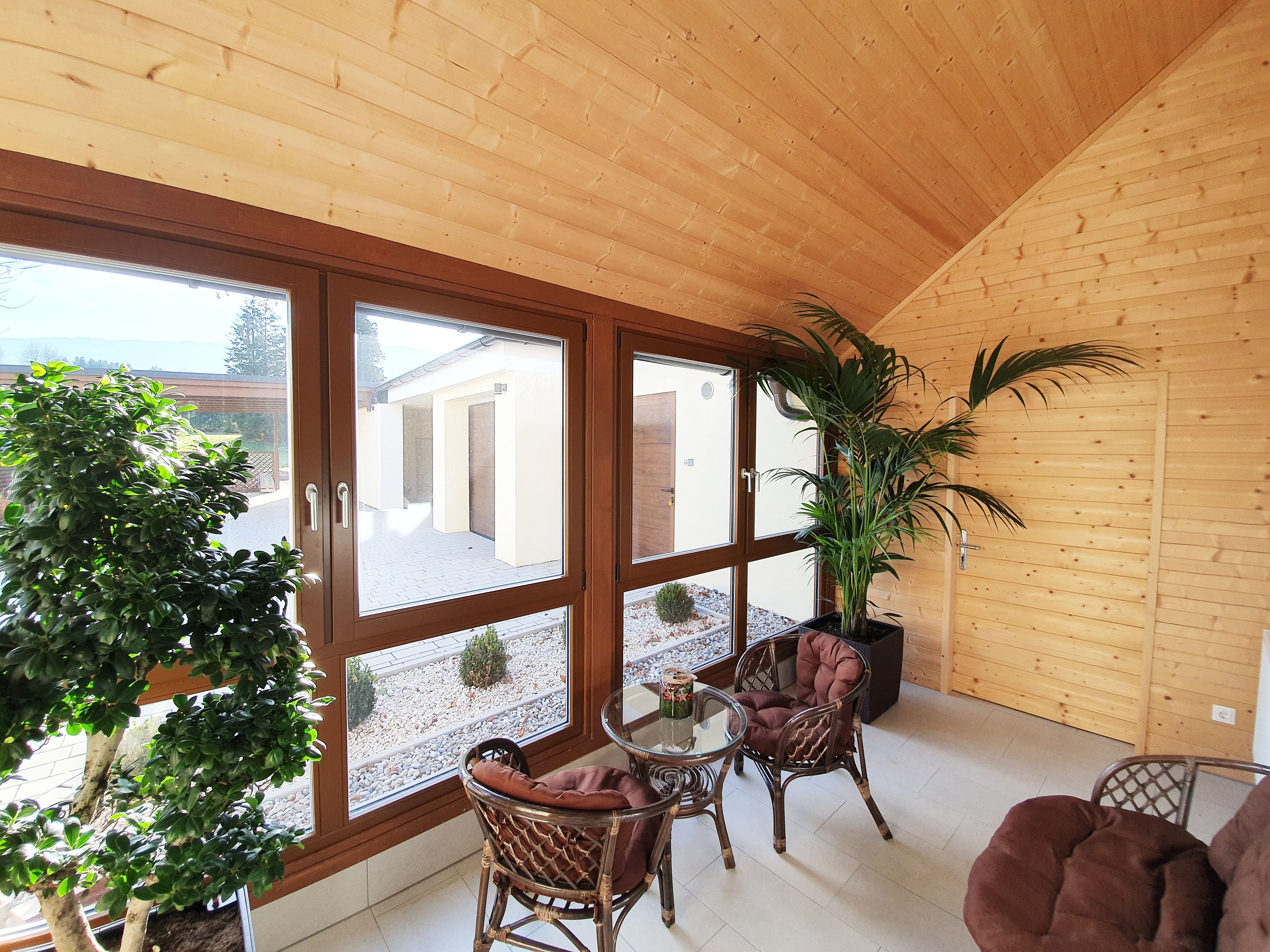 Interior view of a veranda with wooden walls, large windows, plants and rattan furniture.