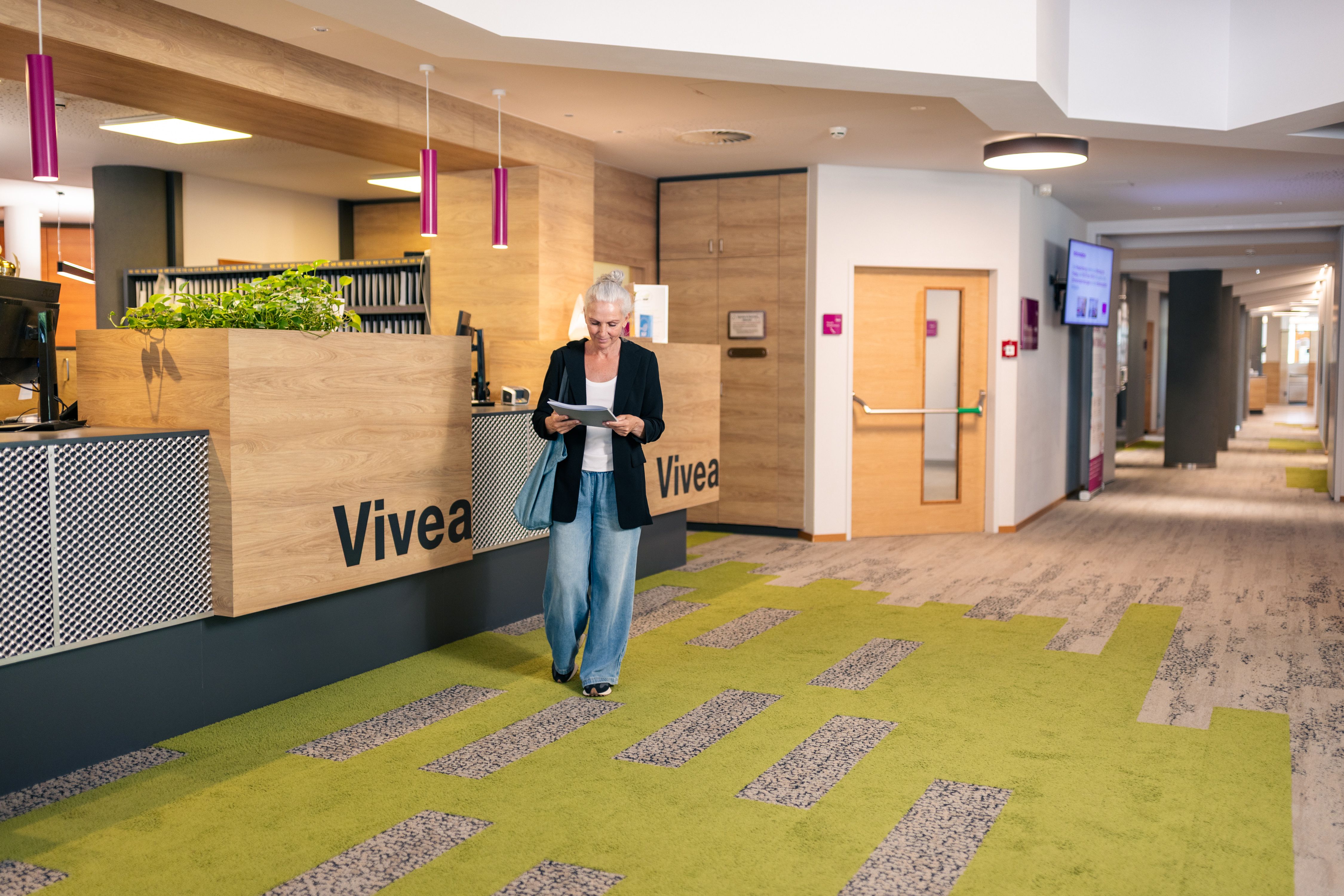 A woman is standing in the lobby of the Vivea 4* Hotel in Bad Schönau, reading a document. The reception area has a modern design featuring wood, plants, and a gray-green carpet.