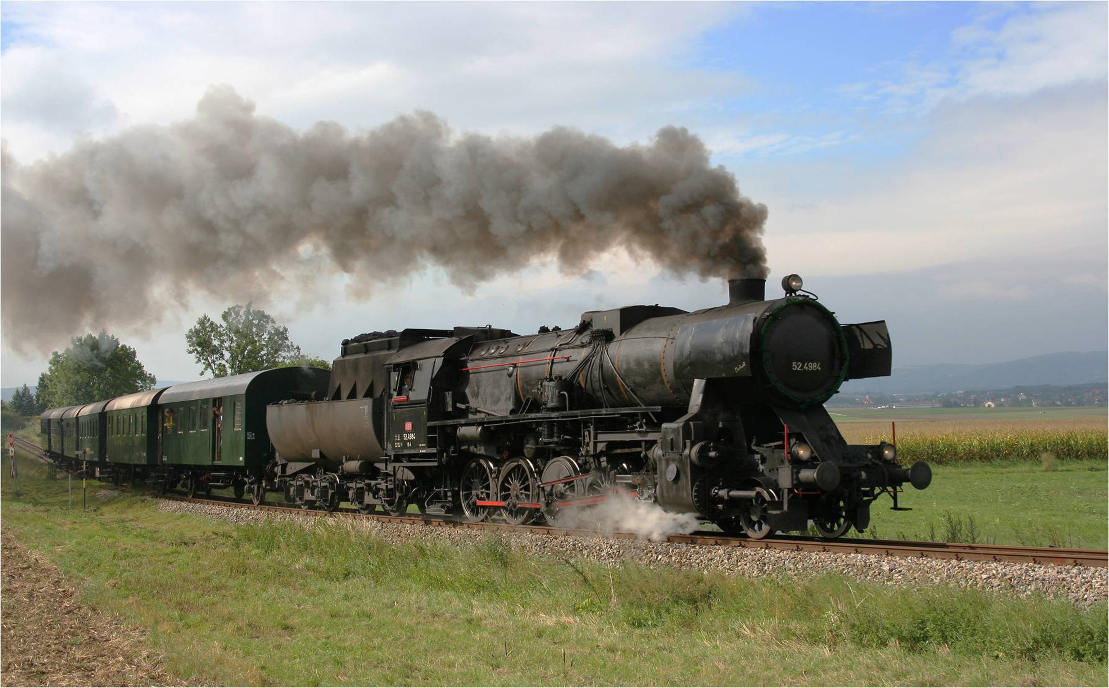 A steaming black steam locomotive pulls green wagons through a rural landscape.