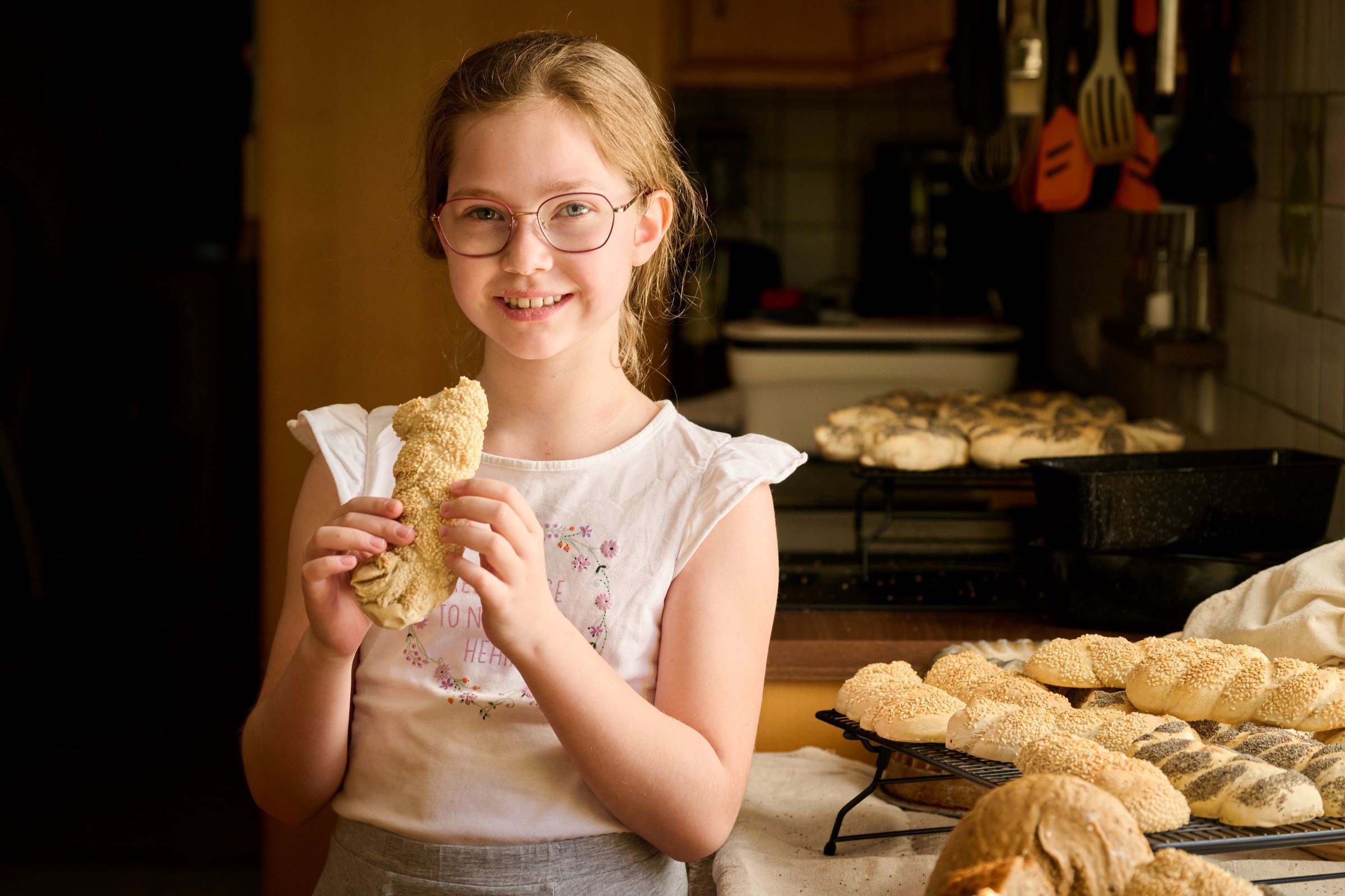 A girl with glasses holds a pastry in her hand and smiles at the camera. Other baked goods can be seen on a table in the background.