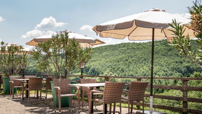 A guest garden with tables, chairs and parasols in front of a green hilly landscape.