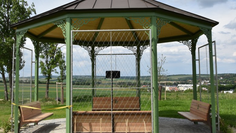 A pavilion with a green metal frame and wooden roof stands on a paved square in a rural setting.
