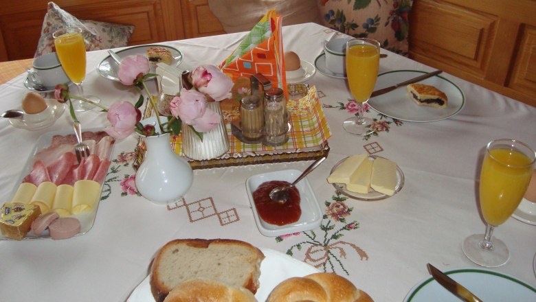 A laid breakfast table with bread rolls, cold cuts, cheese, jam, orange juice and flowers.