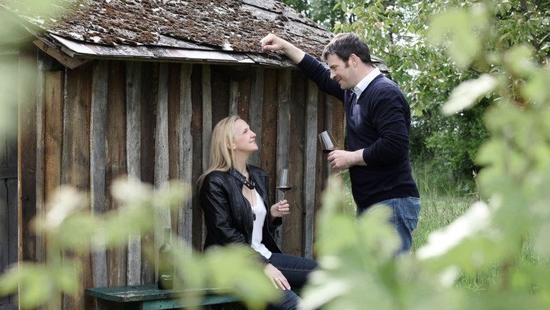 Two people with wine glasses in front of a wooden hut in the countryside.