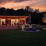 A modern chalet with terrace and sun loungers at sunset, surrounded by trees and an illuminated tree in the garden.