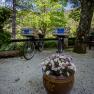 Two bicycles with baskets and "Landsitz Oberhof" signs stand on a gravel path, surrounded by trees and a flower pot with daisies.