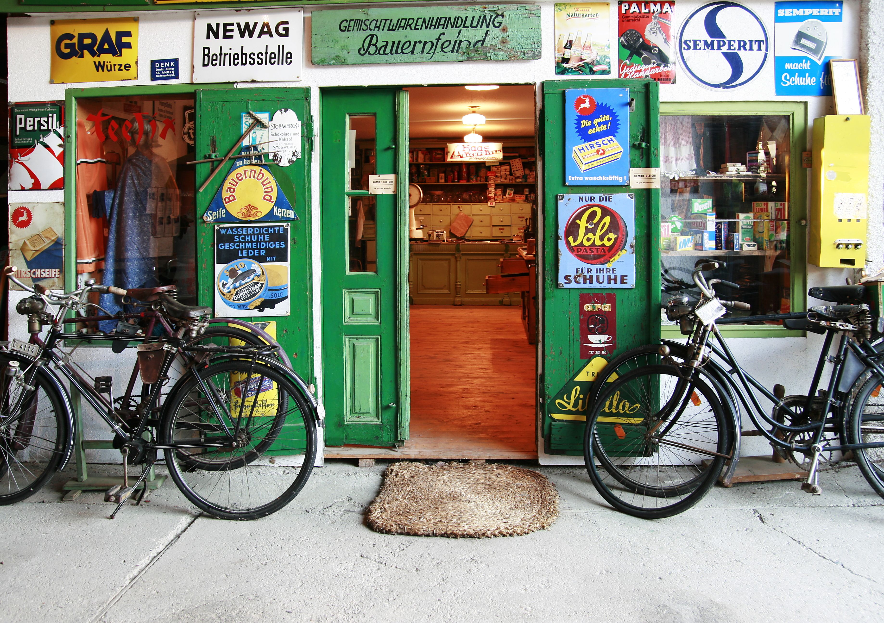 Entrance to an old store with a green door, surrounded by retro advertising signs and two bicycles.