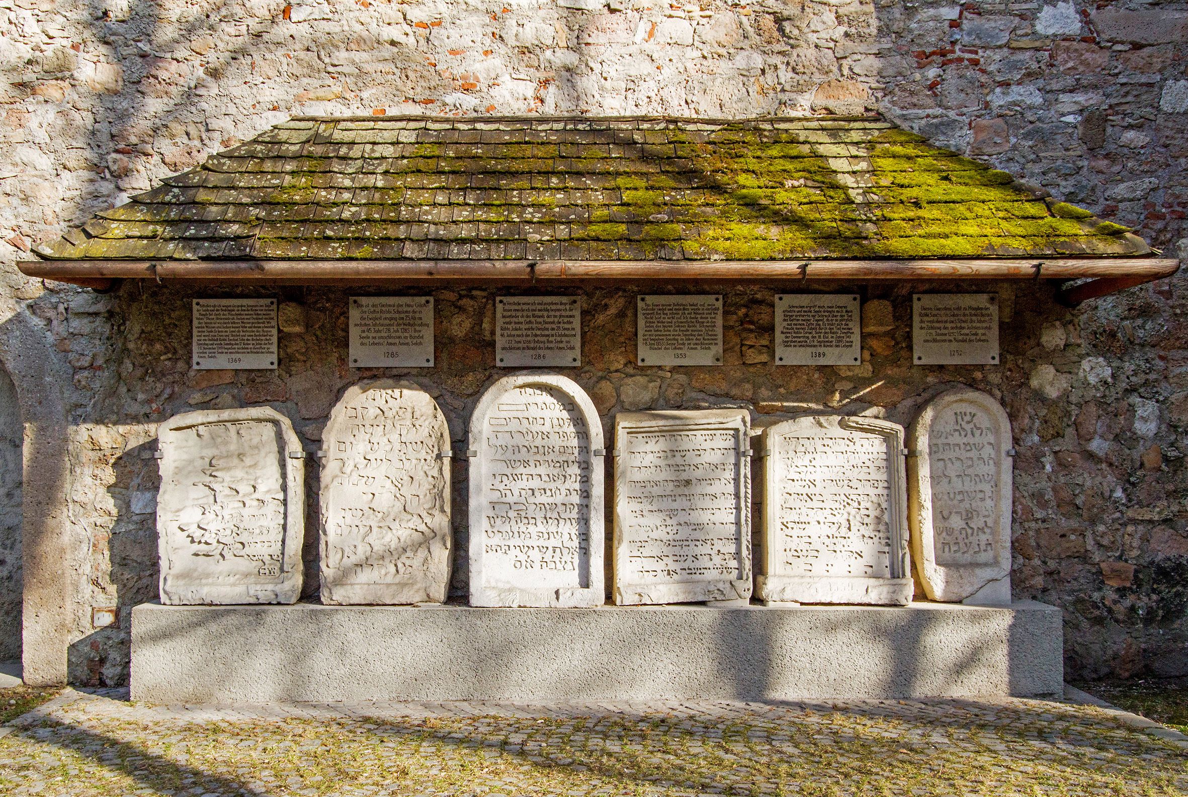 Jewish gravestones on a stone wall with a moss-covered roof.