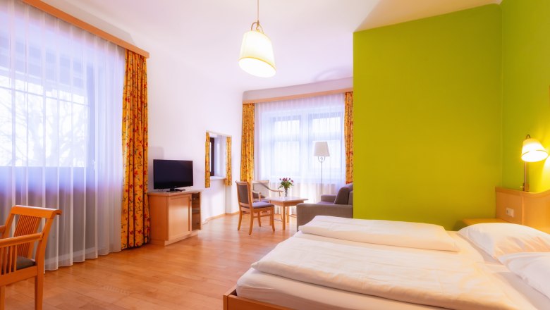 Bright hotel room with double bed, green accent on the wall, TV, seating area and large windows with curtains.