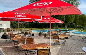 An outdoor pool with red parasols and empty wooden tables on a paved terrace.