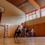 Five teenagers running in a gym with a basketball hoop and handball goal.