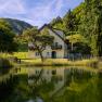 Yellow house with balcony, surrounded by trees, reflected in a pond.