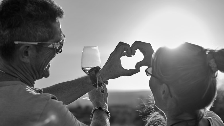 Two people form a heart with their hands in the sunset, one is holding a wine glass.