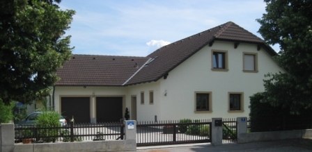 Zelenka family, © Zelenka A modern detached house with a brown roof and light-colored paint, surrounded by trees and a fence.