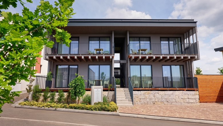 Modern two-storey apartment complex with balconies and staircase, surrounded by plants and trees.