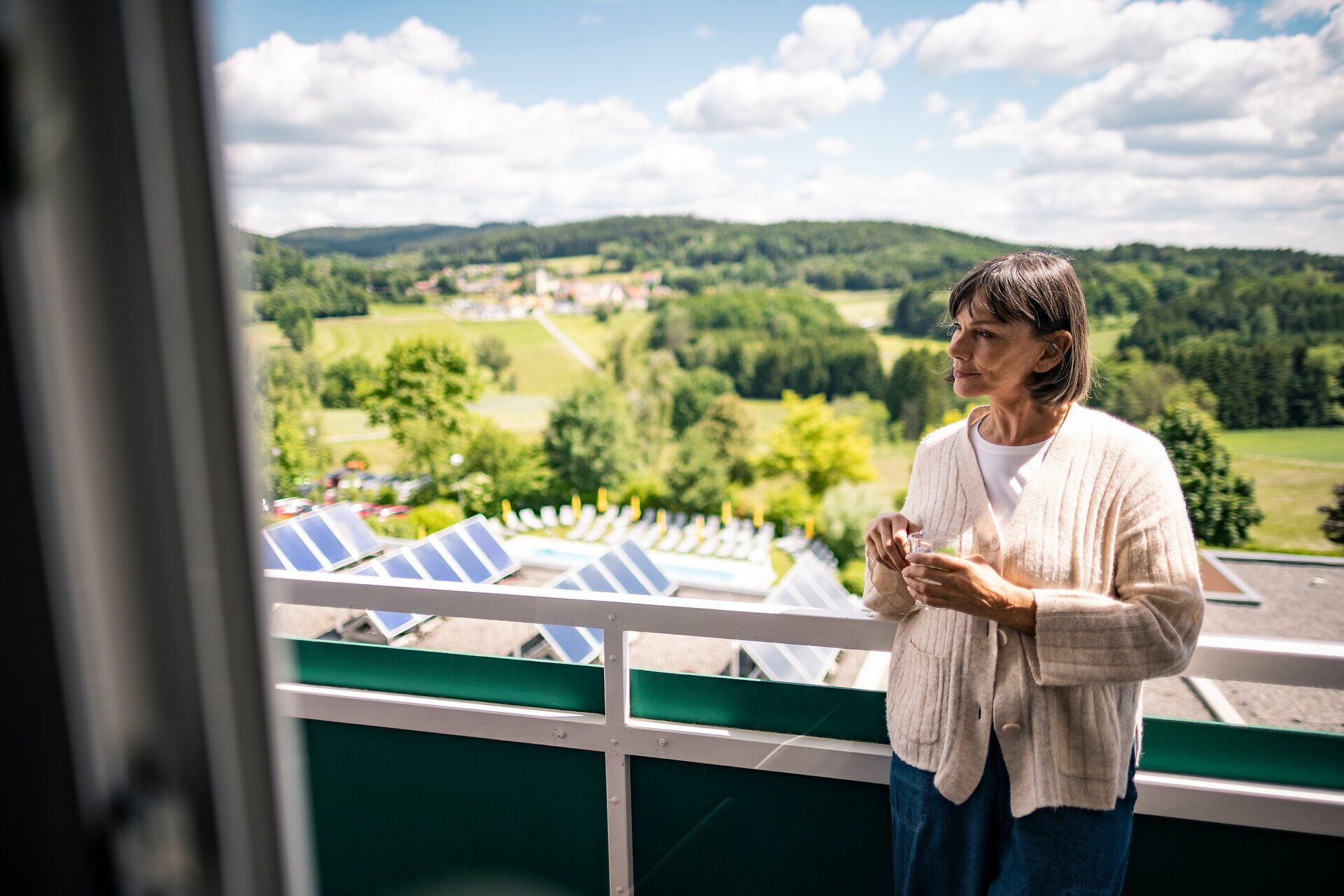 A person holding a cup on the balcony of the Moorheilbad Harbach Health and Rehabilitation Center in the Waldviertel, looking out over the green rolling hills.