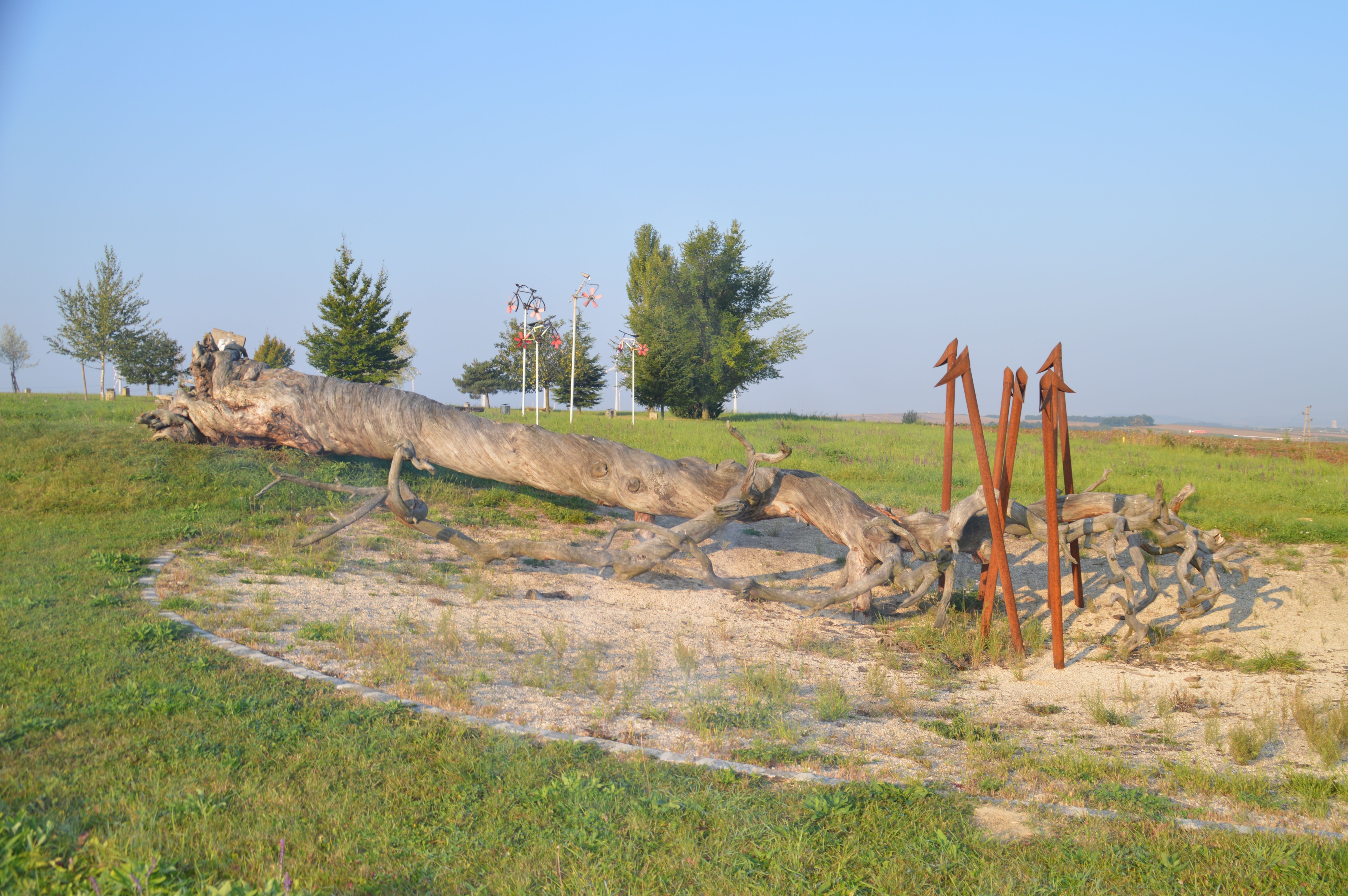 A large, fallen tree trunk lies on a meadow, surrounded by metal sculptures and trees in the background.