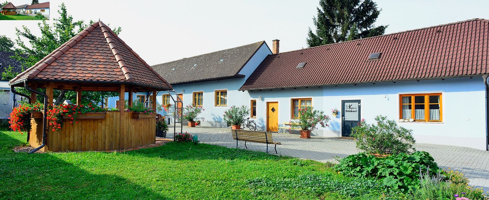 An idyllic courtyard with a pavilion and a blue building with red roofs, surrounded by plants.