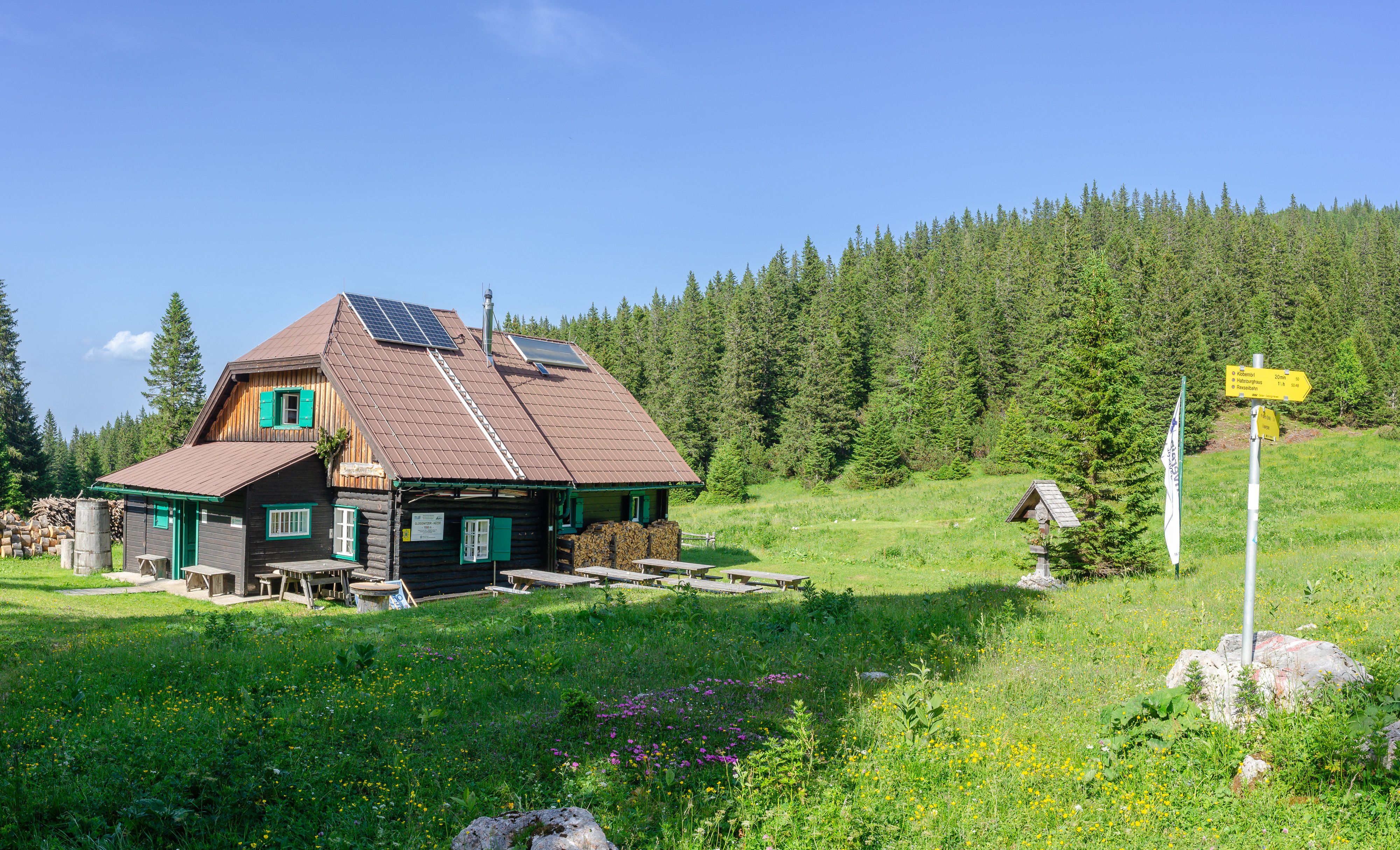 A mountain cabin with solar panels on the roof, surrounded by green meadows and woods.
