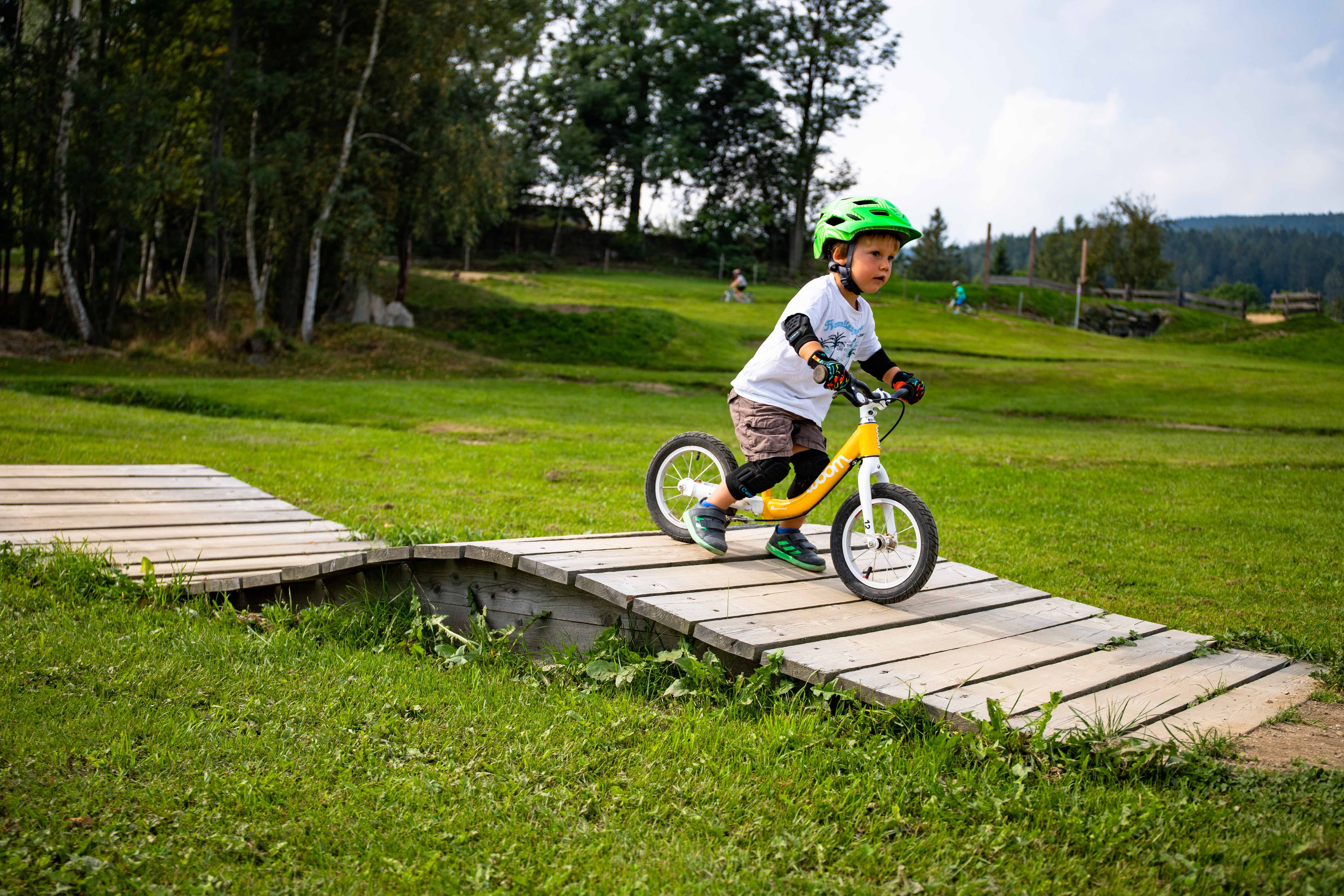 A child with a green helmet rides a balance bike over a wooden ramp in the Mini Bikepark Wexl Trails.
