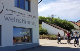 Three people on bicycles ride past a building with the sign 'Weingut - Heuriger - Dworzak weinstimmig'.