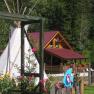 A wooden house with a red roof, a tepee and colorful flowers in the foreground, surrounded by trees.