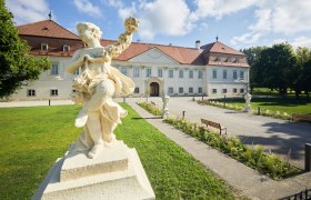 Baroque statue in front of a historic building with a red roof and well-tended garden.