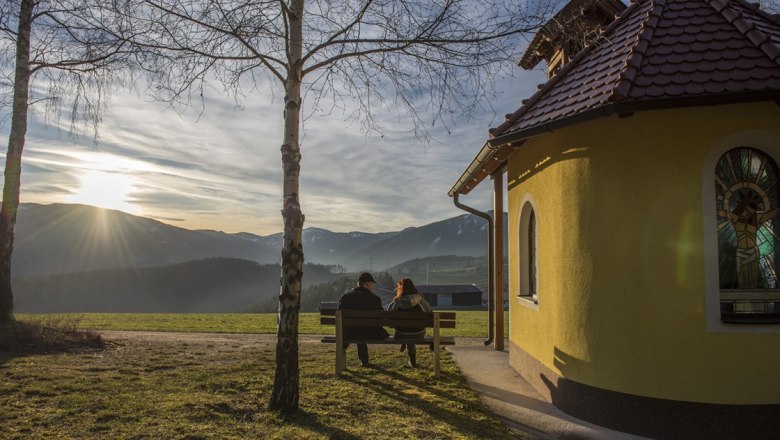 Two people sit on a bench next to a small chapel with a view of the mountains at sunset.