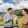 Two men clink glasses of wine, with vineyards and a church tower in the background.
